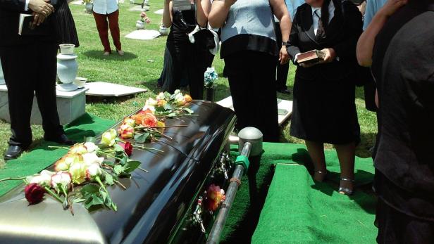 Trauernde Menschen stehen um einen schwarzen Sarg mit Blumen auf einem Friedhof